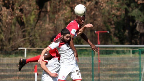 Zwei Fußballspieler kämpfen um den Ball, während ein dritter Spieler im Hintergrund zuschaut. Der Ball ist in der Luft.