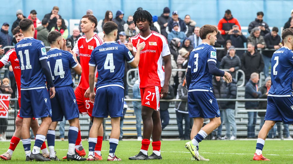 Spieler beider Mannschaften stehen auf dem Spielfeld, einige in roten und andere in blauen Trikots, im Moment der Begrüßung.