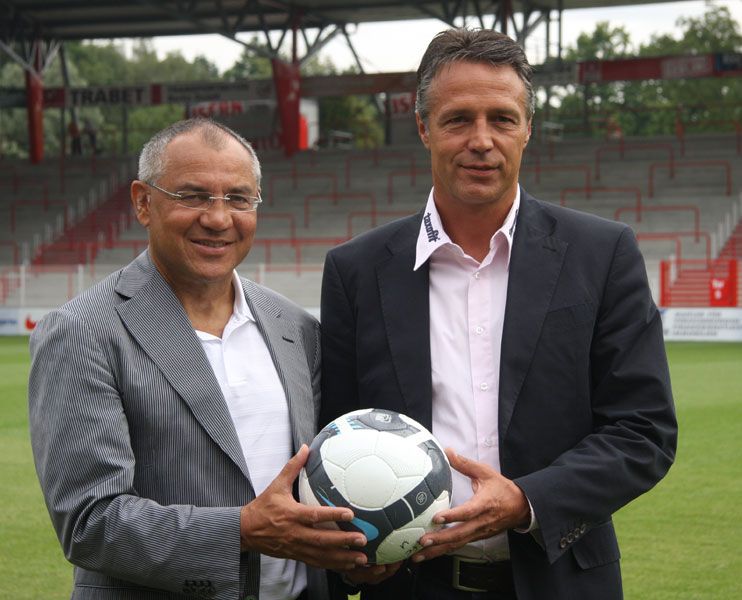 Zwei Männer in Anzügen halten einen Fußball in einem Stadion mit leeren Tribünen im Hintergrund.