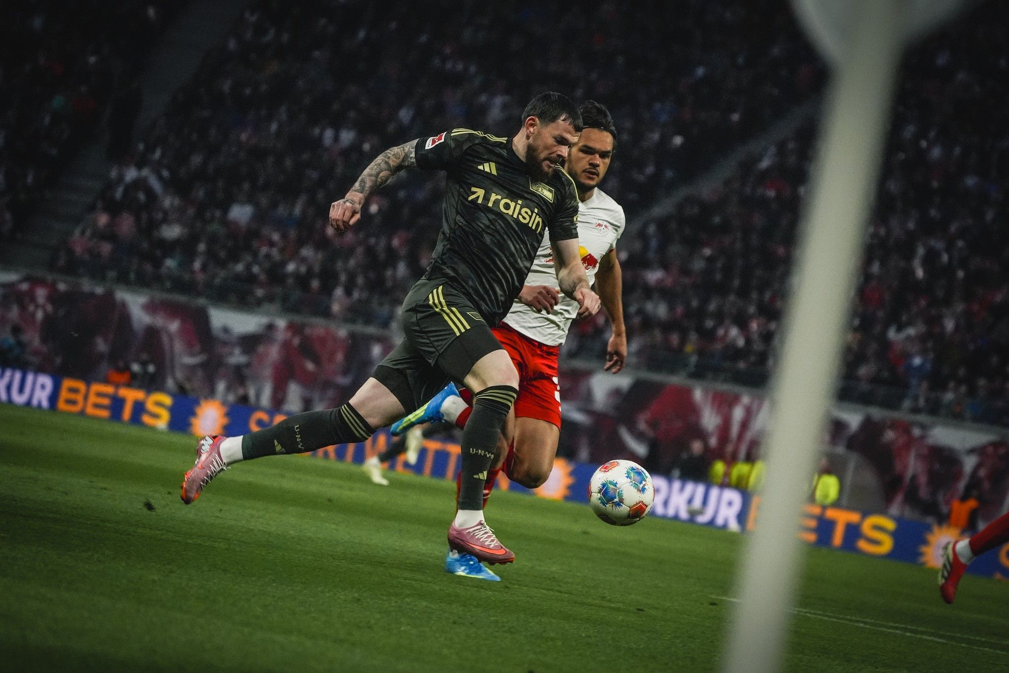 Ein Spieler in schwarzem Trikot läuft mit dem Ball, während ein Gegner im weißen Trikot ihn verfolgt, auf einem Fußballfeld.