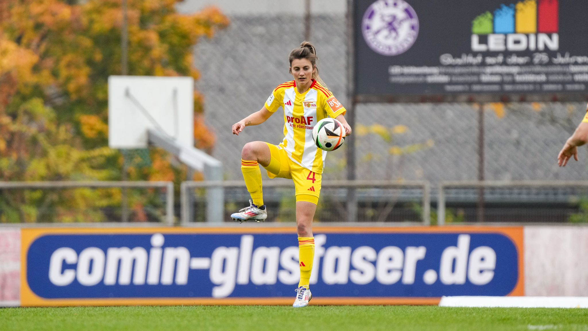 Spielerin in gelb-rot gestreiftem Trikot kontrolliert einen Fußball auf dem Platz. Herbstliche Bäume im Hintergrund.