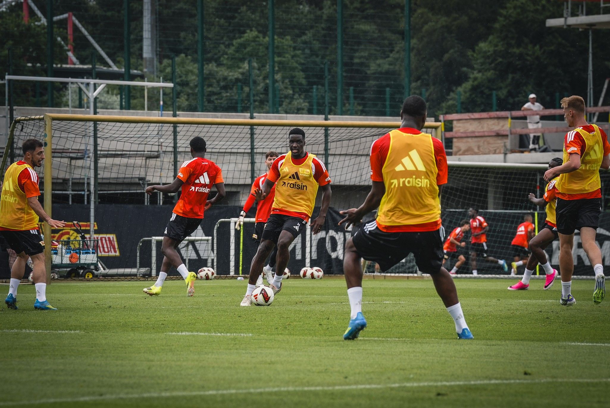 Fußballspiel im Training mit Spielern in orangefarbener und gelber Kleidung auf einem grünen Feld.