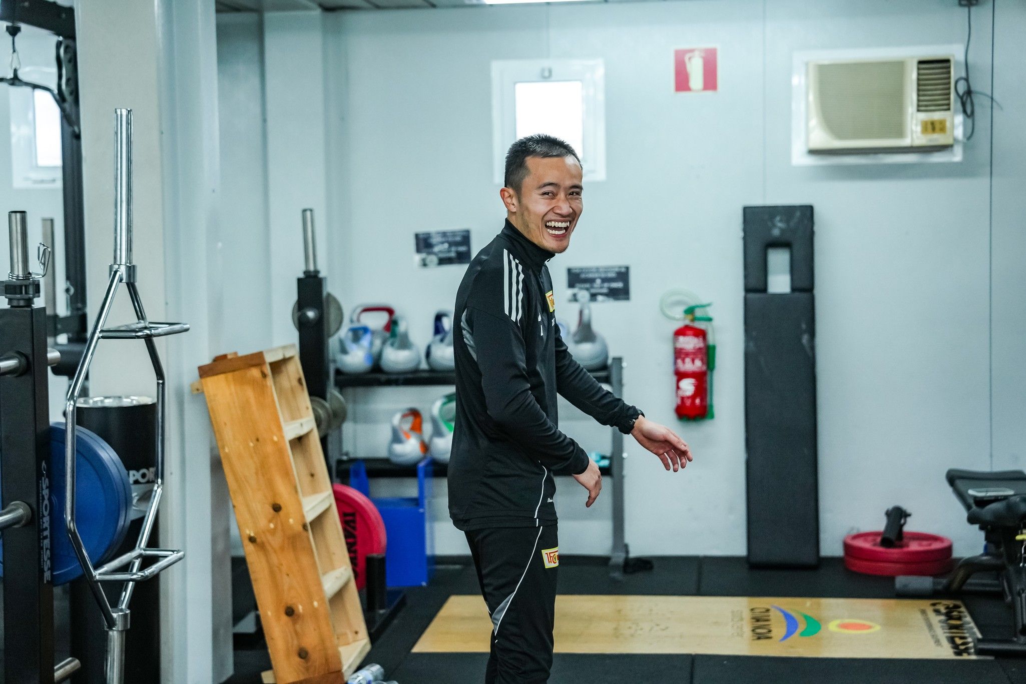 Lächelnder Mann in Sportkleidung steht in einem Fitnessstudio, umgeben von Trainingsgeräten und Zubehör.