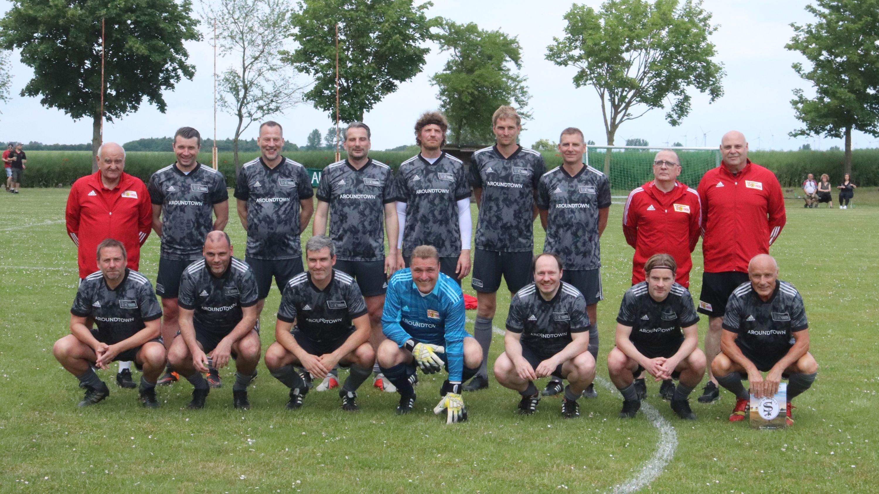 Mannschaftsbild einer Fußballmannschaft in schwarz-grauen Trikots, auf einem Rasenplatz, umgeben von Bäumen.