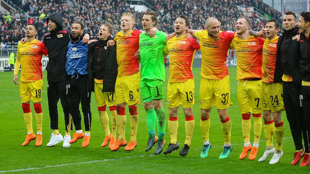 A football team in yellow-red jerseys is cheering in a line, surrounded by a cheering crowd in the background.