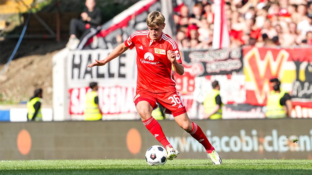 A player in a red jersey dribbles the ball on the field while fans in the background cheer and wave flags.