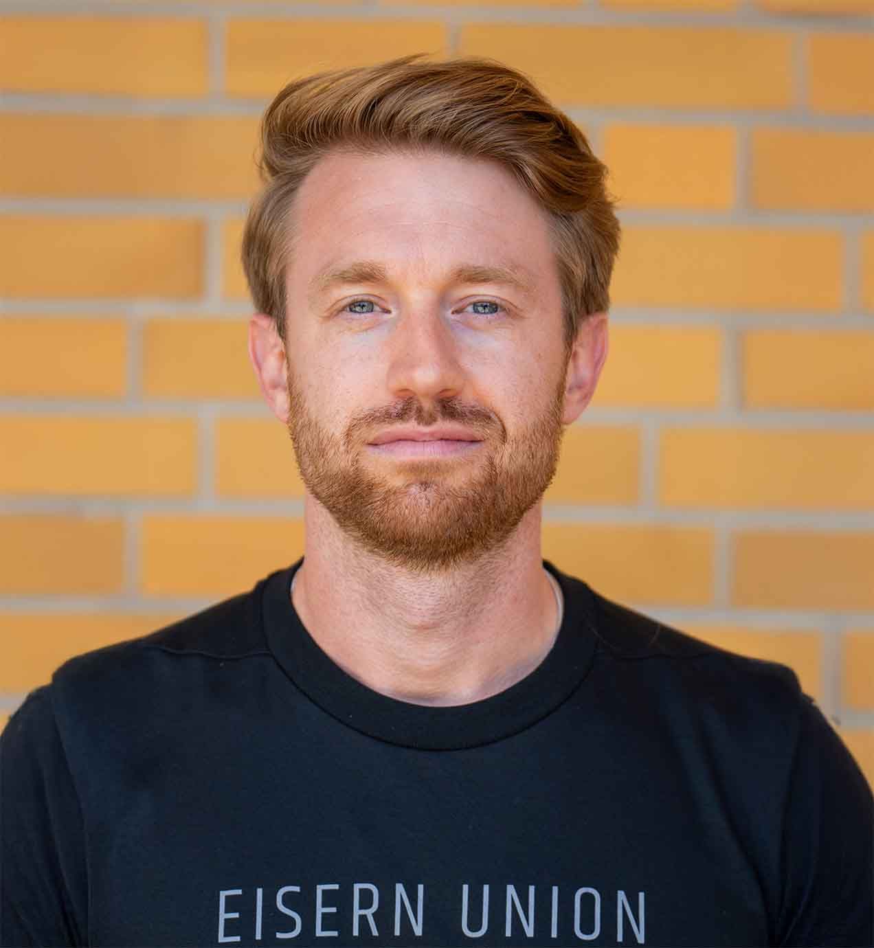 Ein Mann mit blonden Haaren und Bart, trägt ein schwarzes T-Shirt, steht vor einer Backsteinwand.