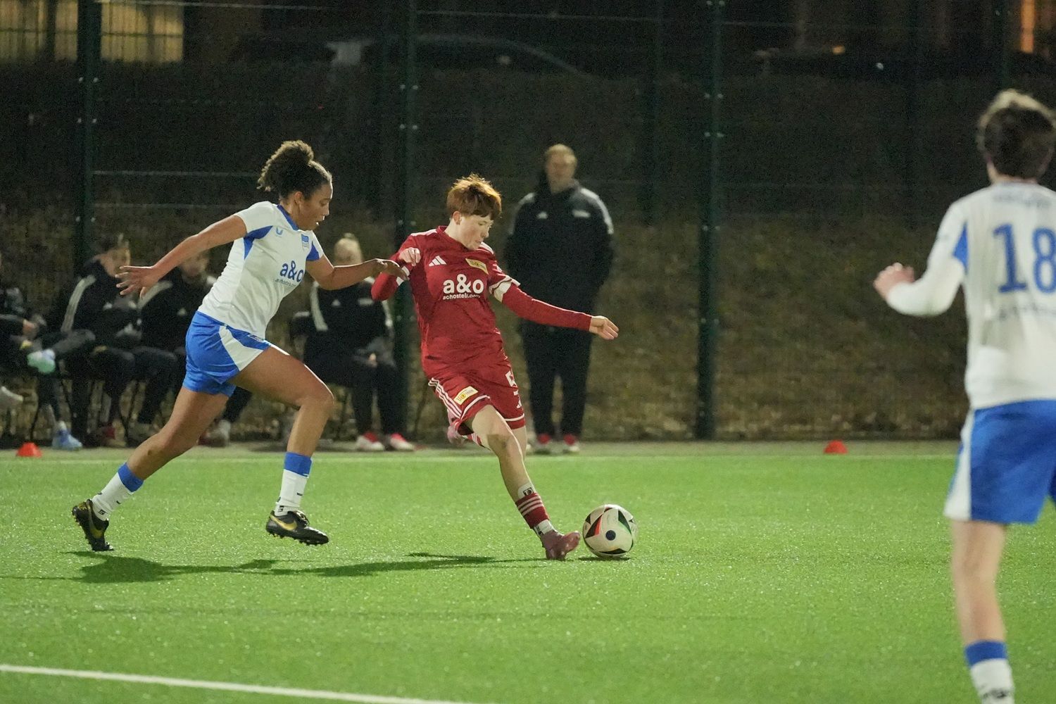 Ein junger Fußballspieler in rotem Trikot schießt den Ball, während eine Spielerin in blauem Trikot ihn verfolgt.