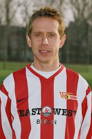 Ein Fußballspieler steht in einem gestreiften Trikot mit dem Vereinslogo und einem Sponsorenschriftzug vor unscharfem Hintergrund.