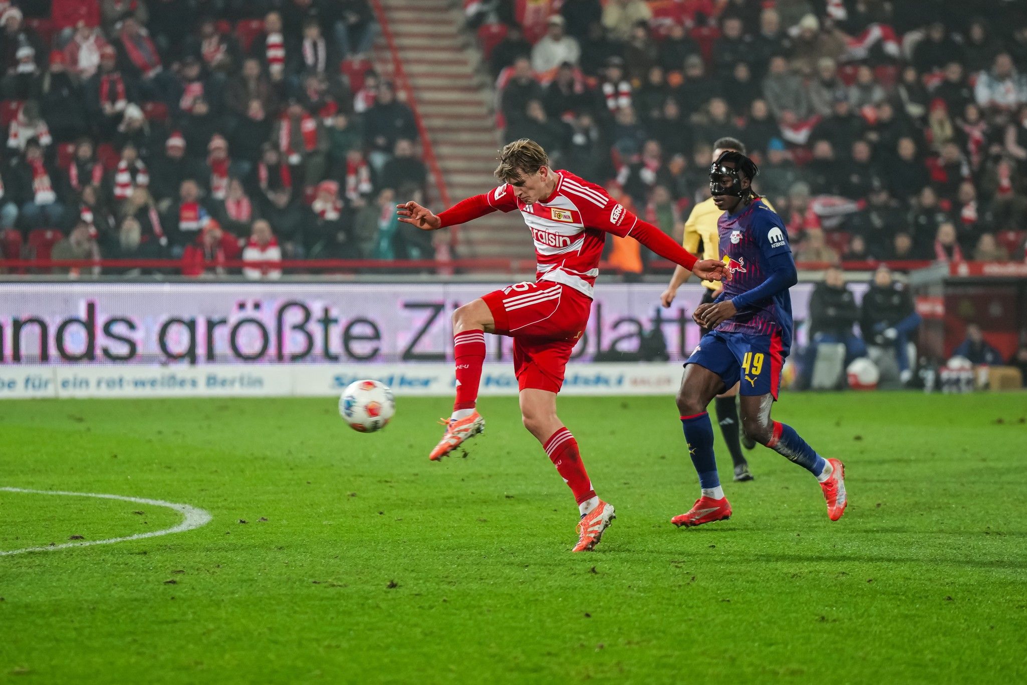 Fußballspiel mit einem Spieler in rotem Trikot, der den Ball mit dem rechten Fuß passt, während ein Gegner in blauem Trikot dabei ist.
