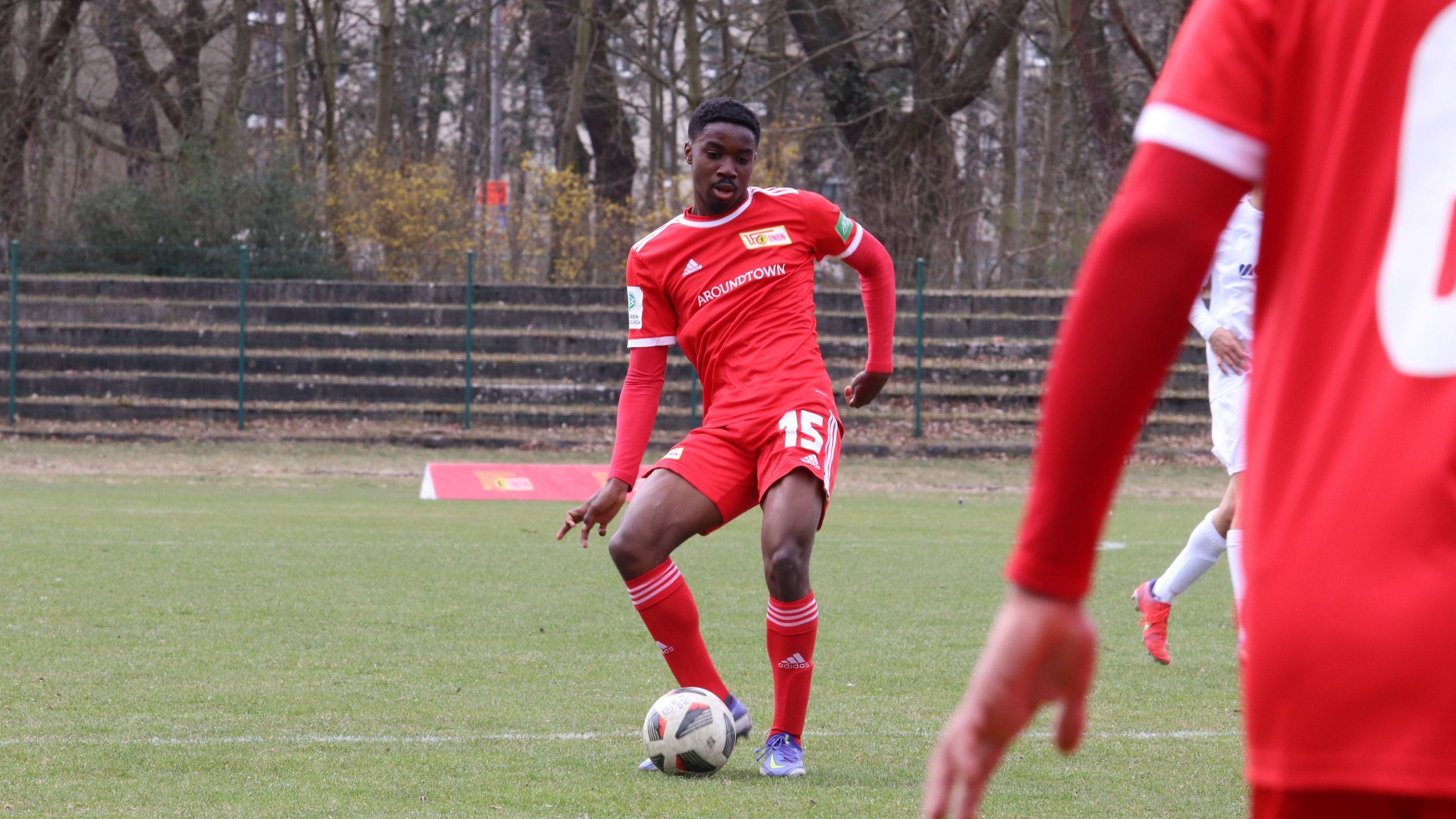 Ein Fußballspieler in rotem Trikot dribbelt den Ball auf einem Rasenplatz, umgeben von Bäumen und Zuschauerbänken.
