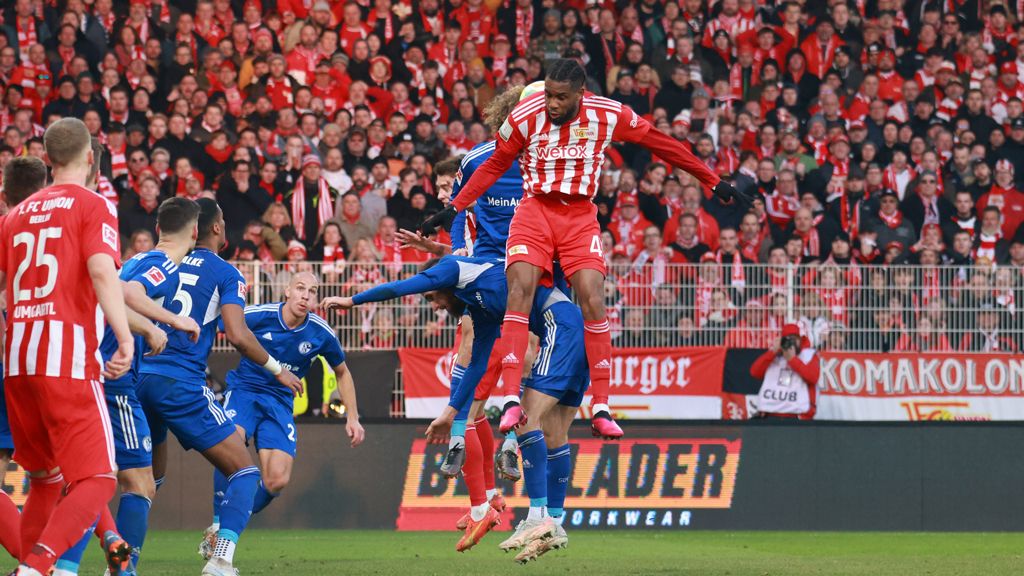 Spielszene aus einem Fußballmatch, Spieler springen zum Kopfball, Fans im Hintergrund jubeln.