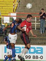 Zwei Fußballspieler springen zum Kopfball, während ein Zuschauer im Hintergrund steht.