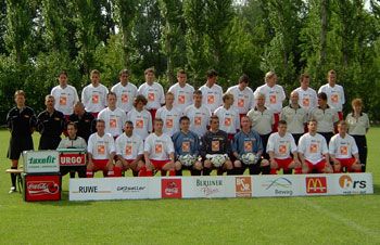 Eine Fußballmannschaft steht auf einem grünen Rasenfeld, gekleidet in weiße Trikots mit Sponsorenlogos, umgeben von Trainer und Betreuern.