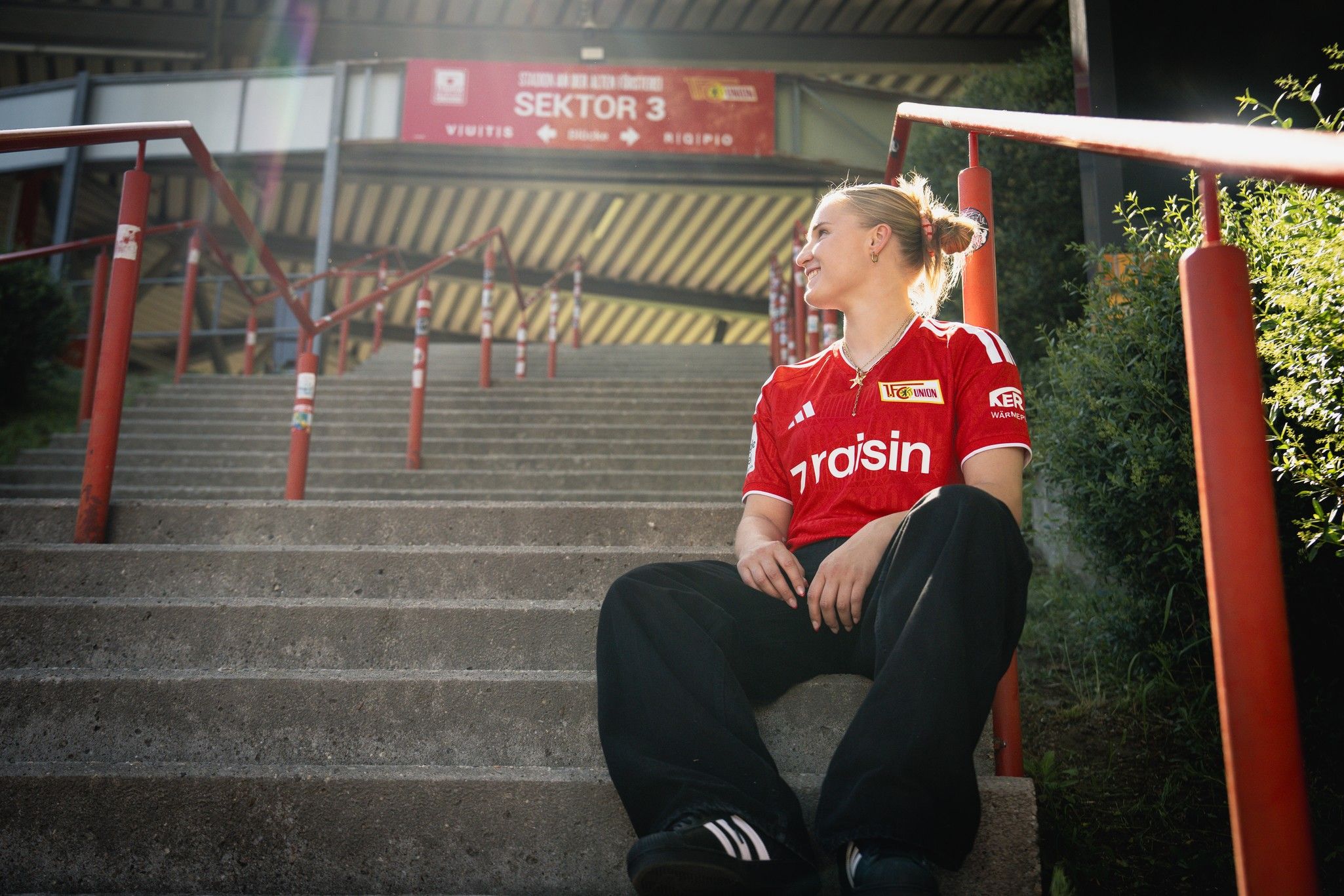 Eine Person in einem roten Sporttrikot sitzt auf einer Treppe und schaut lächelnd nach oben. Im Hintergrund sind Stufen und eine Stadionanzeige.