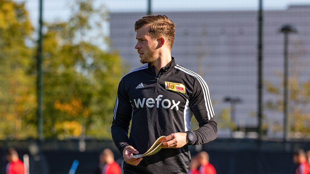 A coach in sportswear is taking notes during a training session on the field. Blurry players are visible in the background.