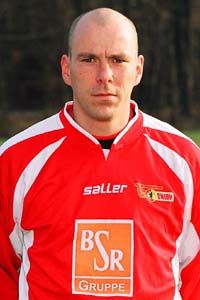 Ein sportlicher Mann trägt ein rotes Fußballtrikot mit dem Logo eines Teams und einem Sponsor. Er hat eine glatte, rasierte Kopfhaut.