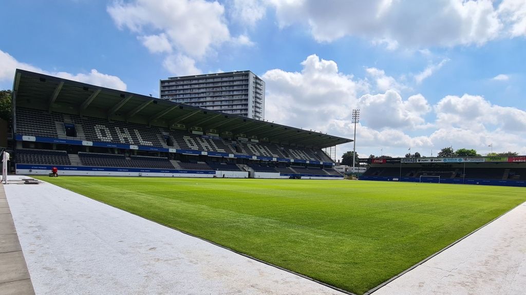 View of a football stadium with green grass, seating, and a tall building in the background under a cloudy sky.