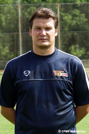 Ein männlicher Sporttrainer steht in einem blauen Trikot auf einem Fußballfeld, im Hintergrund sind Sporteinrichtungen sichtbar.