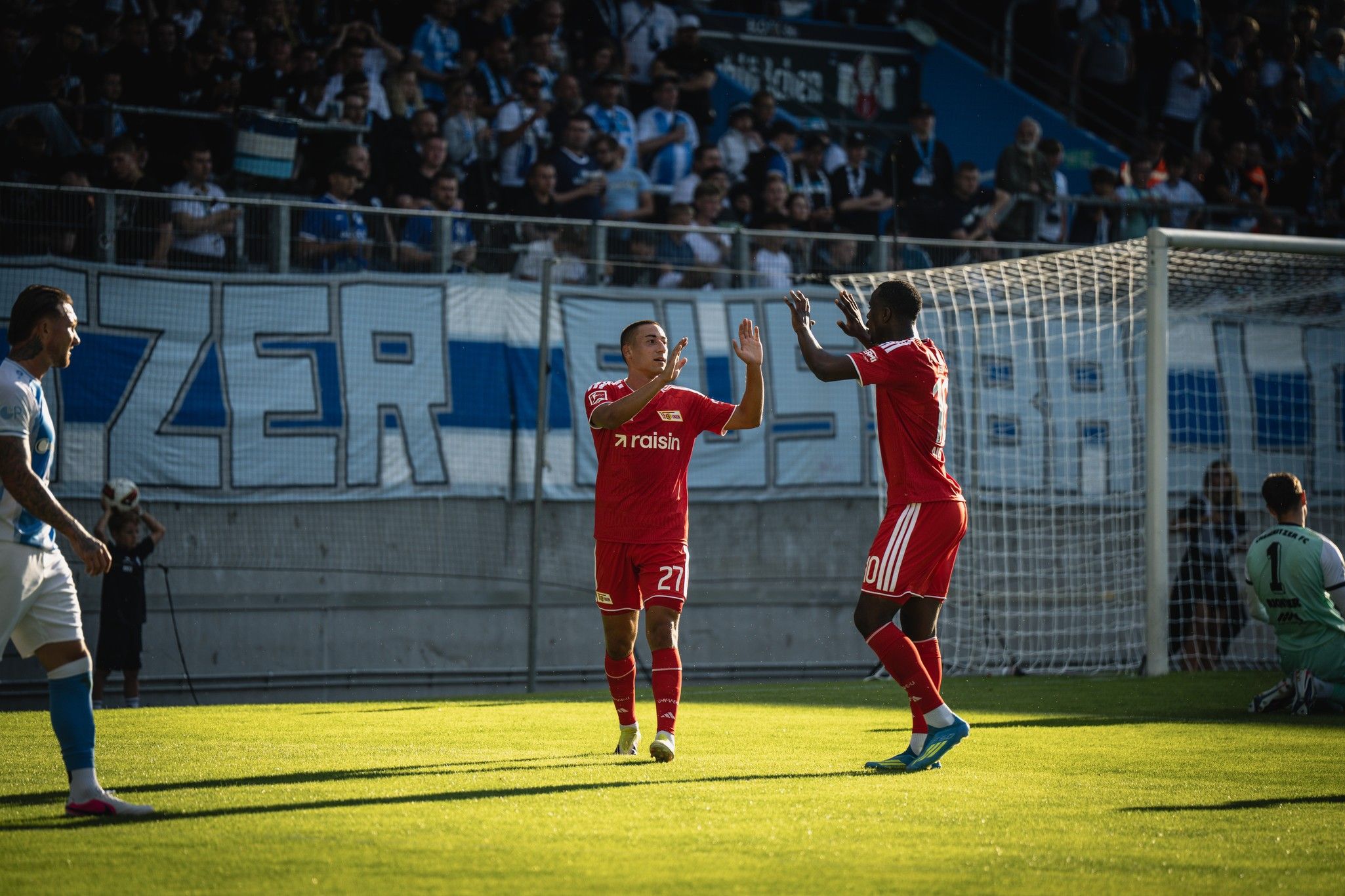Zwei Fußballspieler im roten Trikot feiern einen Spielmoment auf dem Platz, während Zuschauer im Hintergrund jubeln.