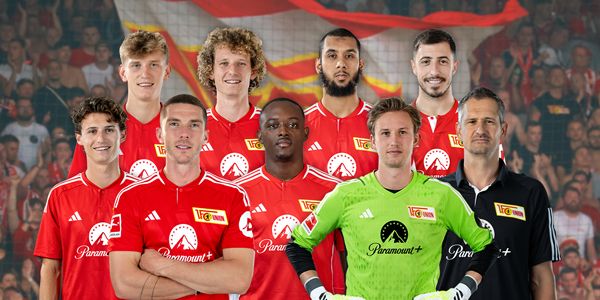 Eine Gruppe von Fußballspielern in roten Trikots steht auf einem Platz, umgeben von Fans im Hintergrund.