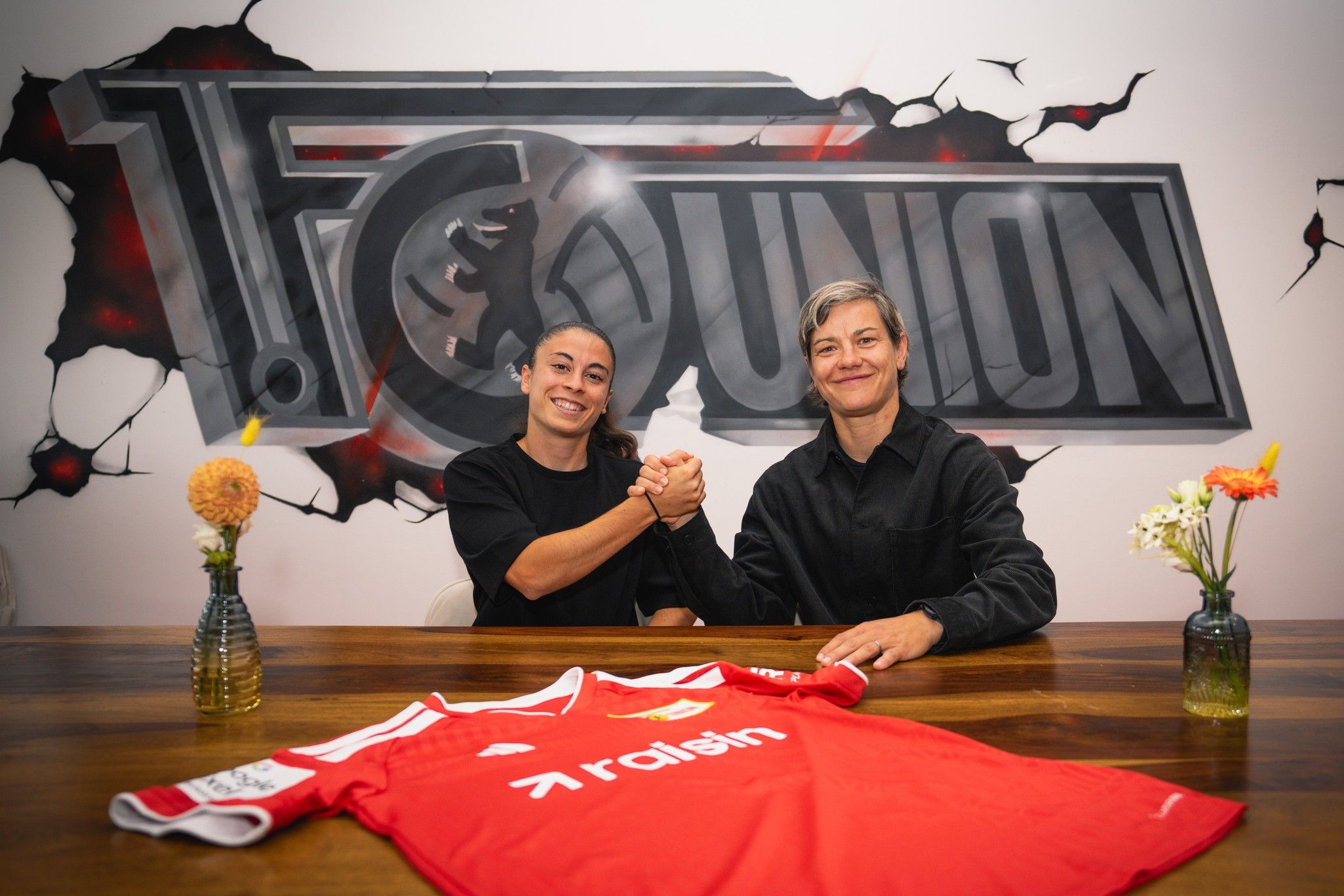 Zwei Personen schütteln sich die Hände vor einer Wand mit dem Logo von 1. FC Union und einem roten Fußballtrikot auf dem Tisch.