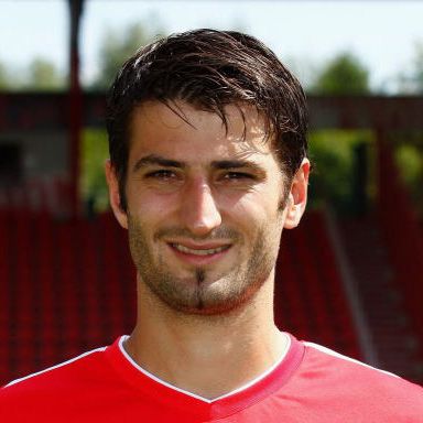 Porträt eines lächelnden Fußballspielers in rotem Trikot, vor einem Stadionhintergrund.