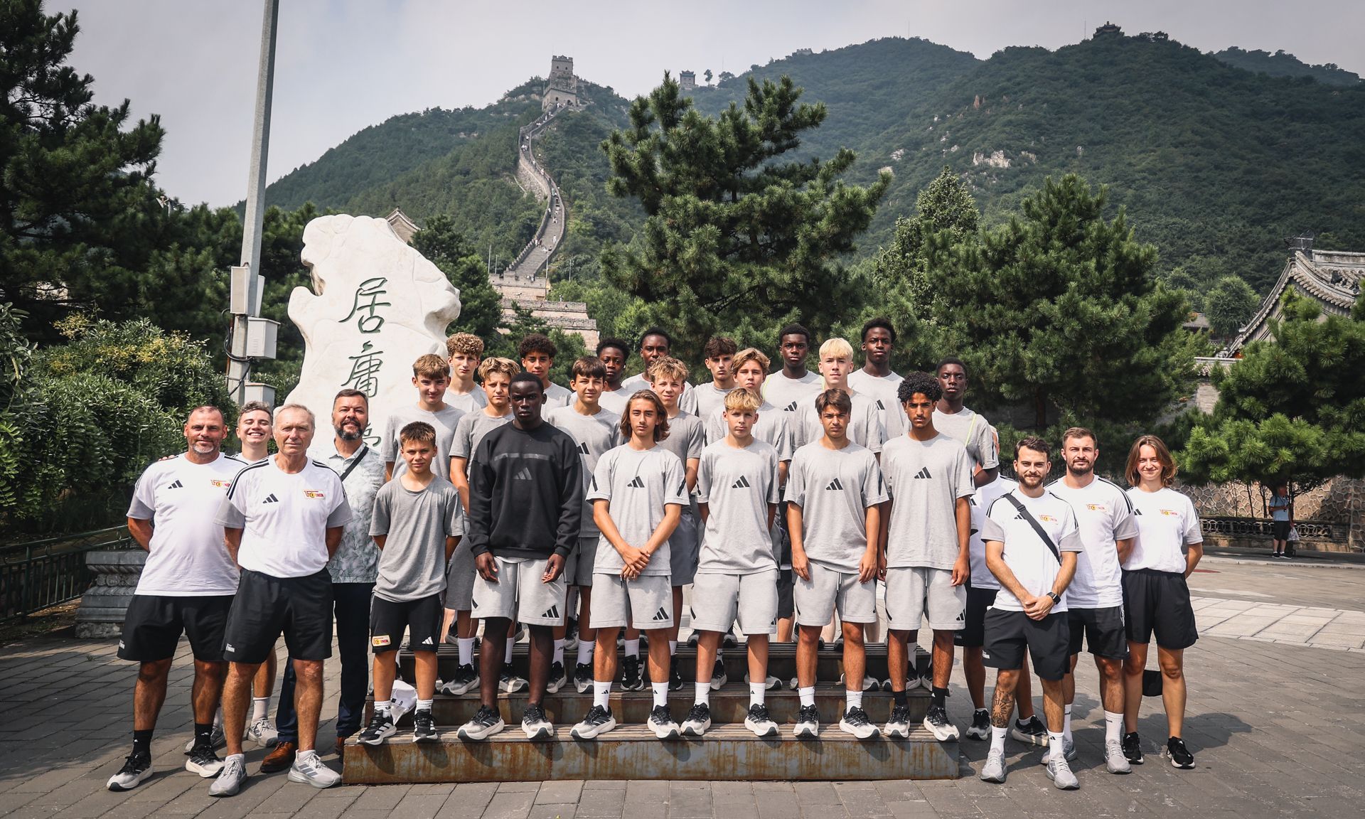 Gruppe von Jugendlichen in grauen Sportoutfits posiert vor der Chinesischen Mauer, umgeben von Bäumen.