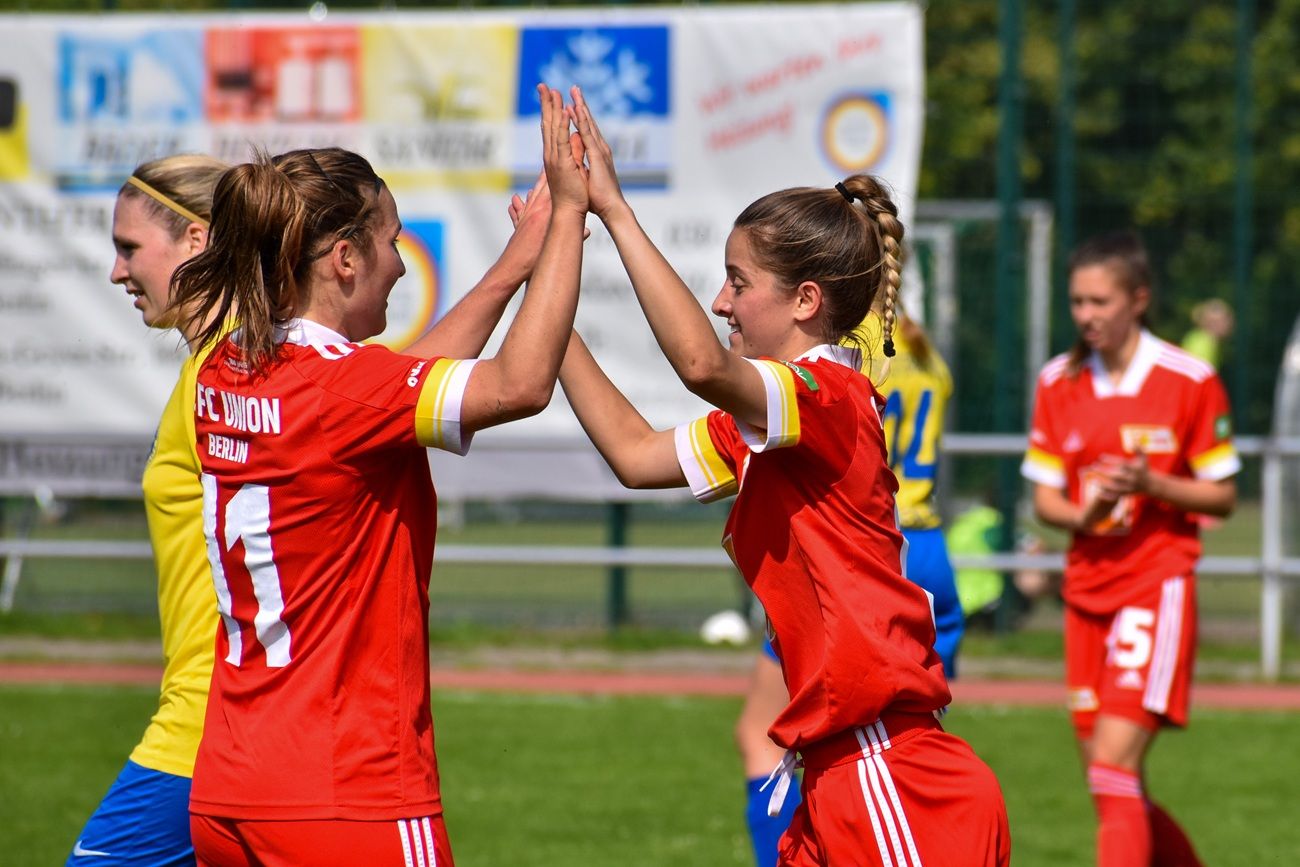 Zwei Spielerinnen in roten Trikots feiern mit einem High-Five auf dem Fußballfeld. Im Hintergrund sind weitere Spielerinnen zu sehen.