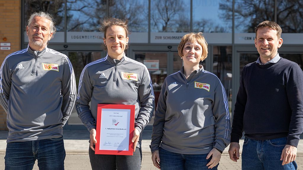 Vier Personen stehen vor einem Gebäude und halten ein Zertifikat in der Hand. Sie tragen graue Sportjacken mit Logos.