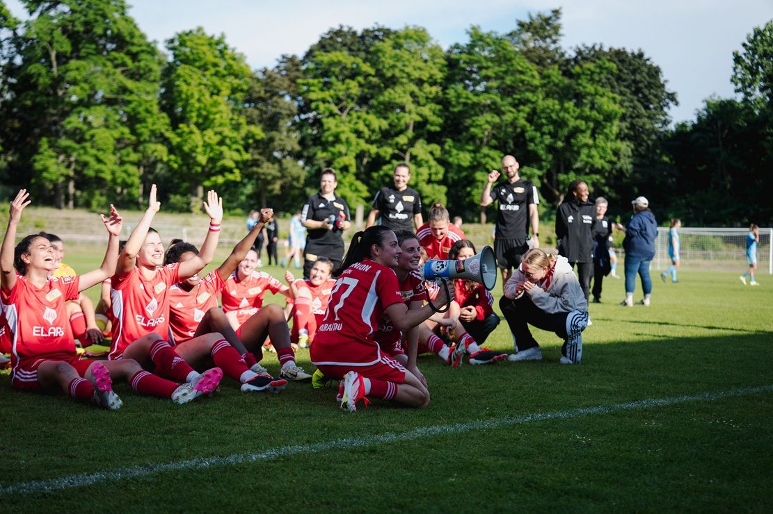 Eine feiernde Fußballmannschaft in roten Trikots sitzt auf dem Rasen, während einige Spieler jubeln und andere lächeln.