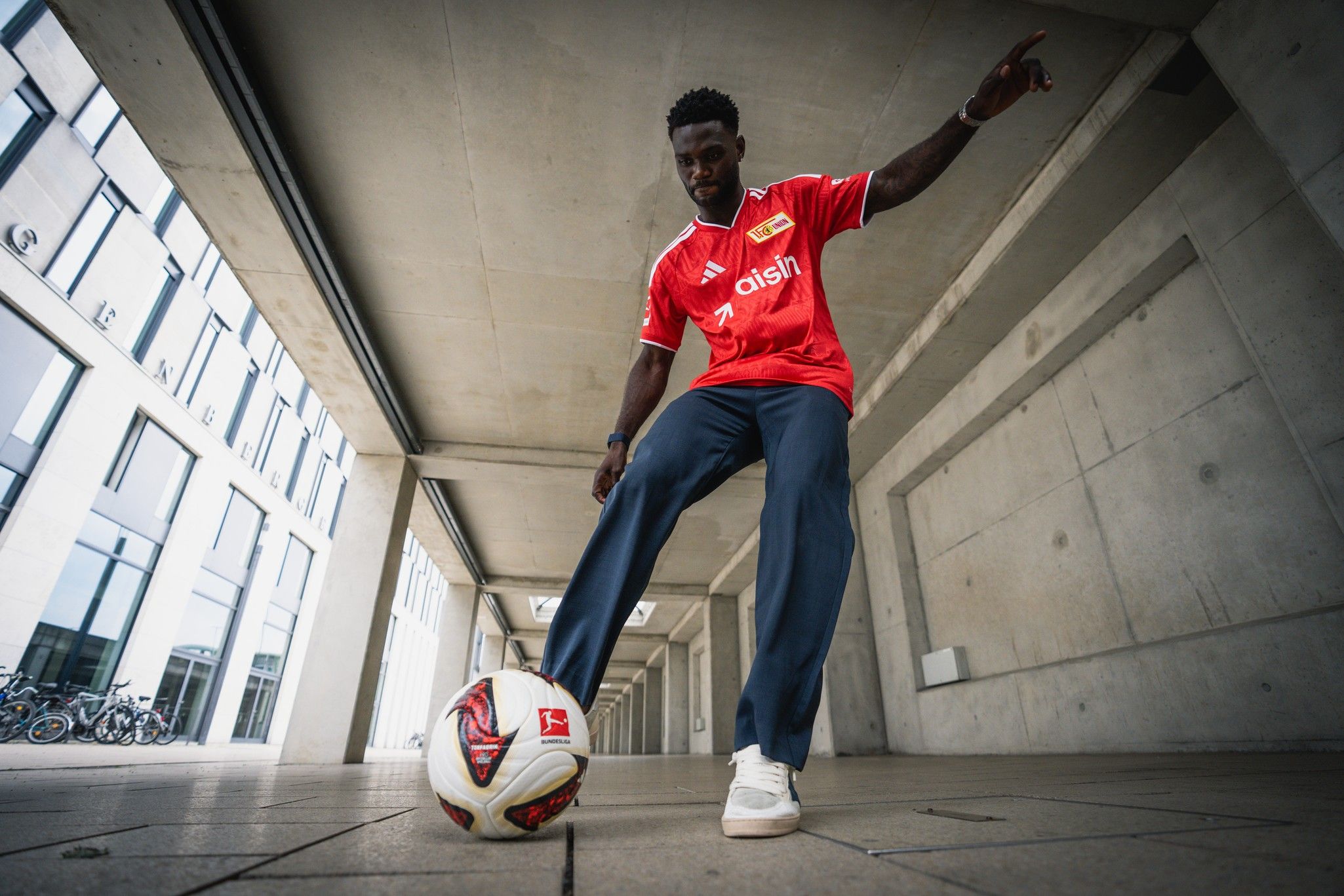 Ein Fußballspieler mit schwarzer Hautfarbe steht in einem modernen Innenhof und dribbelt einen Ball. Er trägt ein rotes Trikot und schwarze Hosen.
