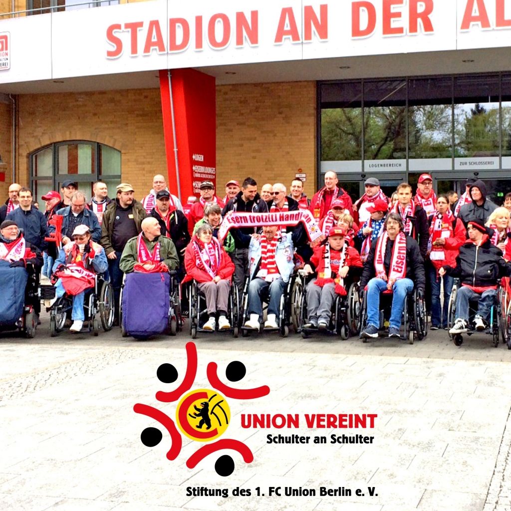 Eine Gruppe von Menschen in Rollstühlen mit Fan-Accessoires posiert vor einem Stadion. 