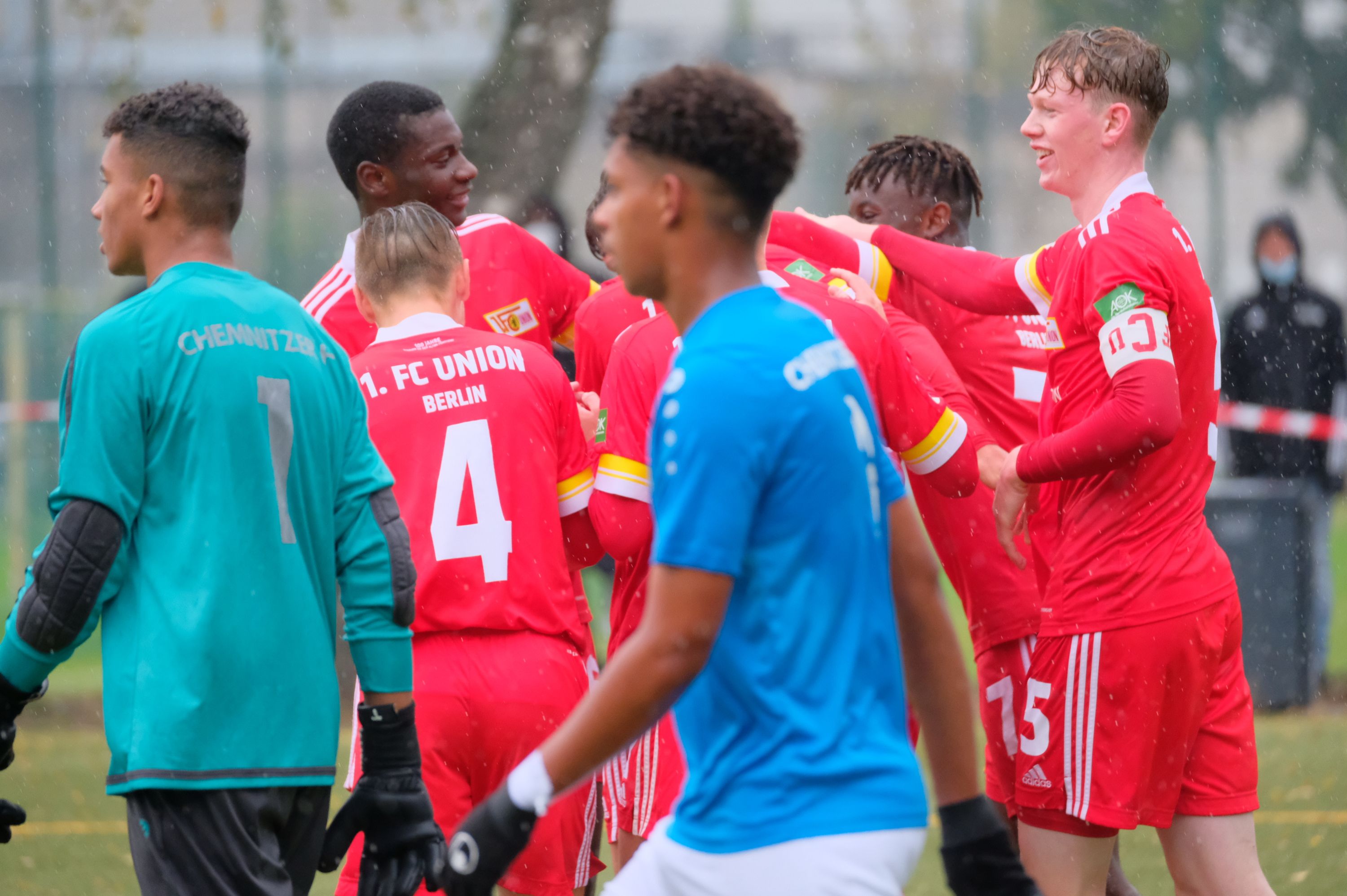 Spieler der Fußballmannschaft in roten Trikots feiern nach einem Tor, während ein Torwart in blauer Uniform im Hintergrund steht.
