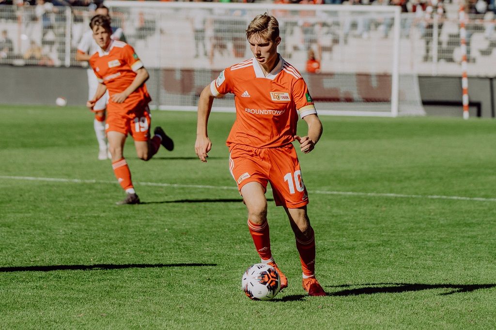 Ein Fußballspieler in orangeem Trikot dribbelt den Ball auf einem grünen Feld, während ein weiterer Spieler im Hintergrund läuft.