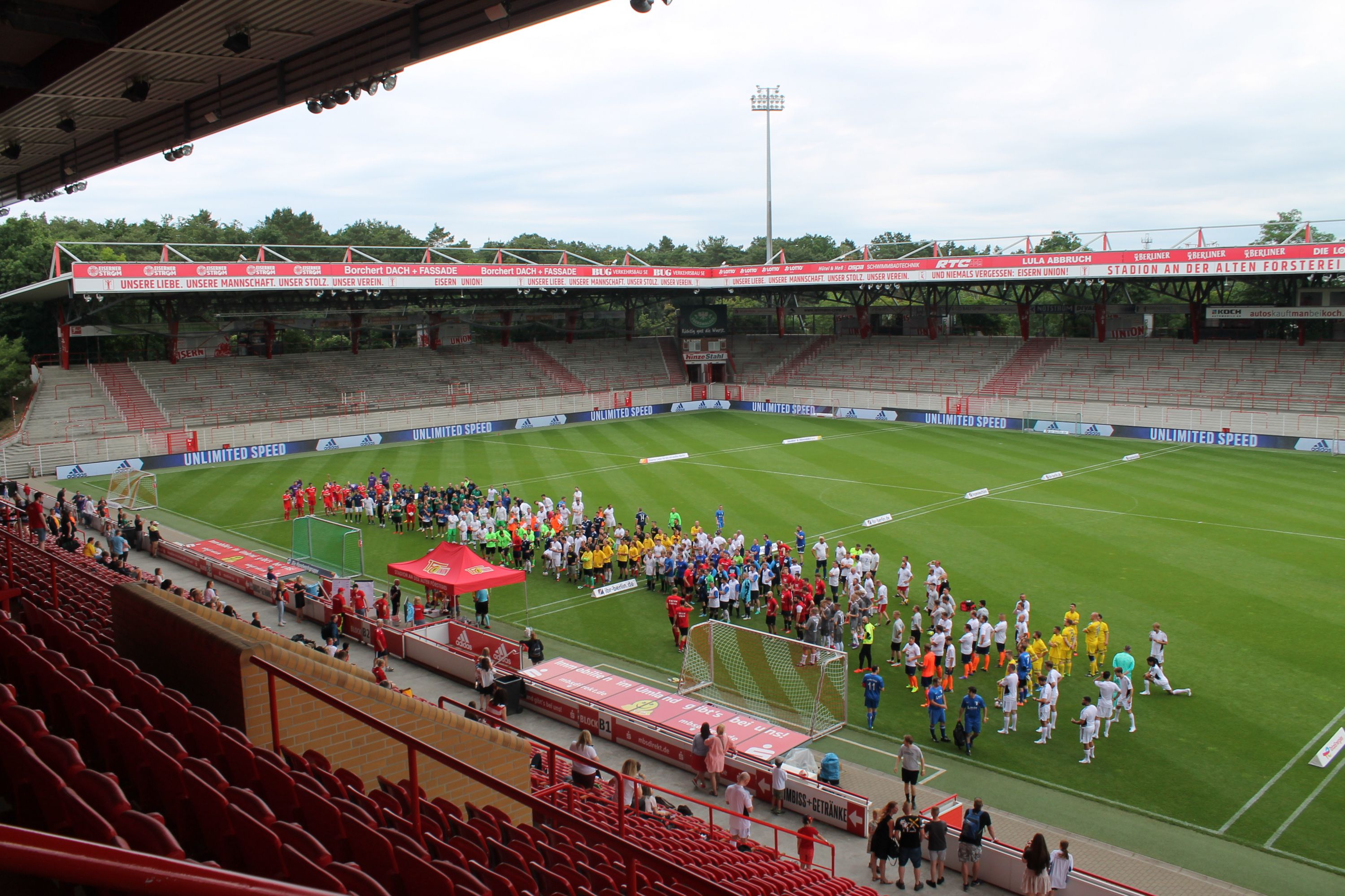 Verschiedene Fußballmannschaften versammeln sich auf dem Spielfeld in einem Stadion mit Sitzplätzen im Hintergrund.