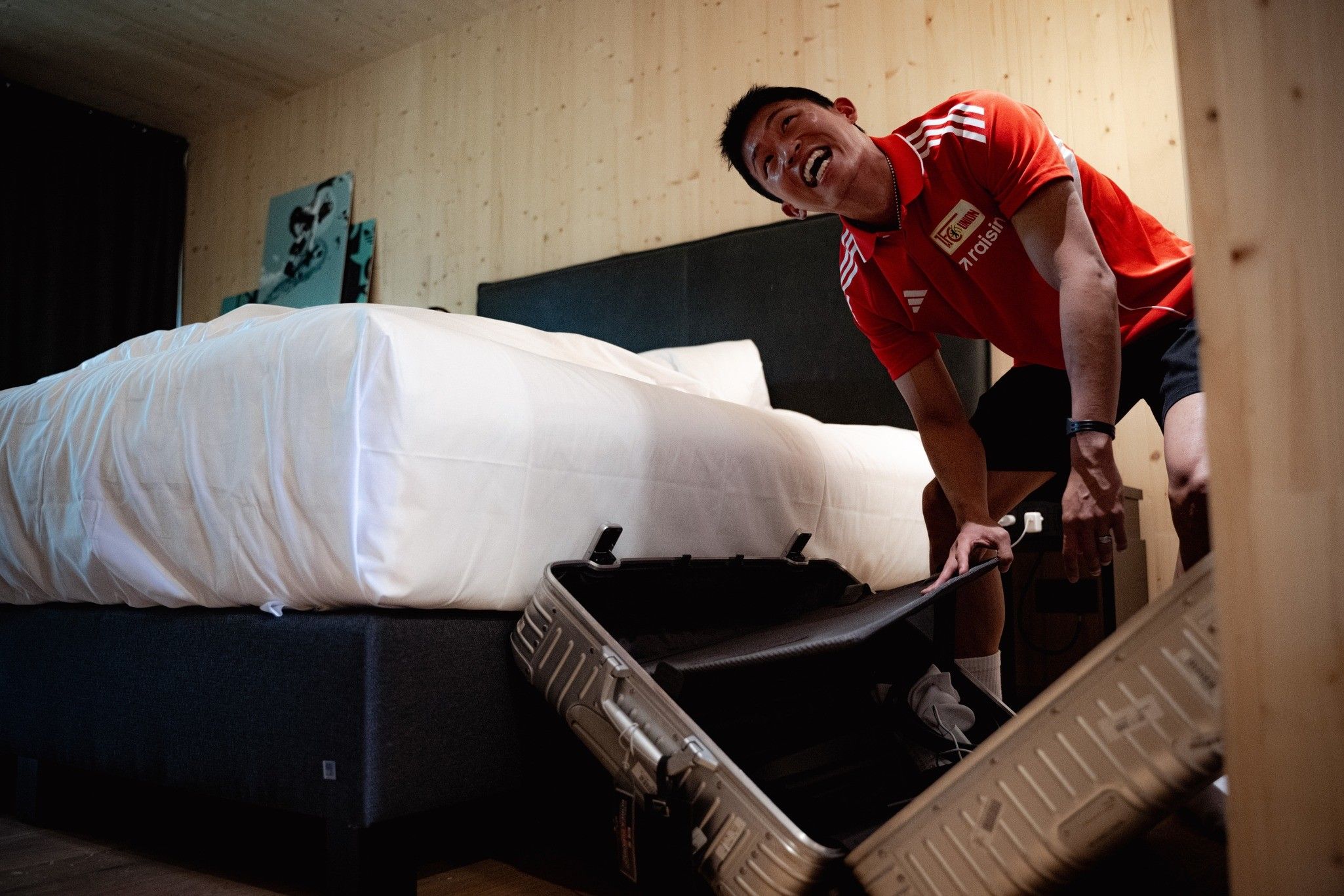 Junger Mann in einem roten Sporttrikot öffnet einen Koffer in einem Hotelzimmer mit einem Bett im Hintergrund.