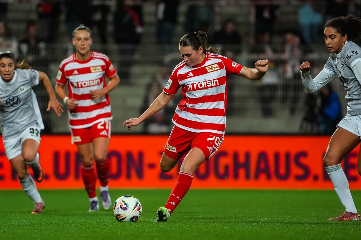 Eine Fußballspielerin in rot-gestreiftem Trikot dribbelt den Ball, während zwei Gegnerinnen in grauen Trikots sie verfolgen.