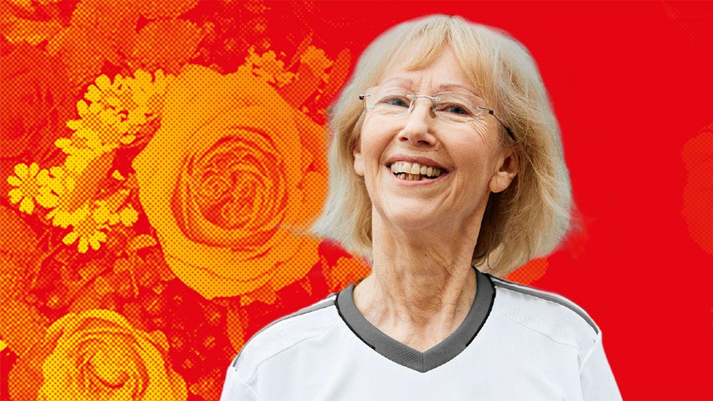 Eine lächelnde ältere Frau mit blonden Haaren, die ein sportliches Oberteil trägt, vor einem roten Hintergrund mit floralen Mustern.