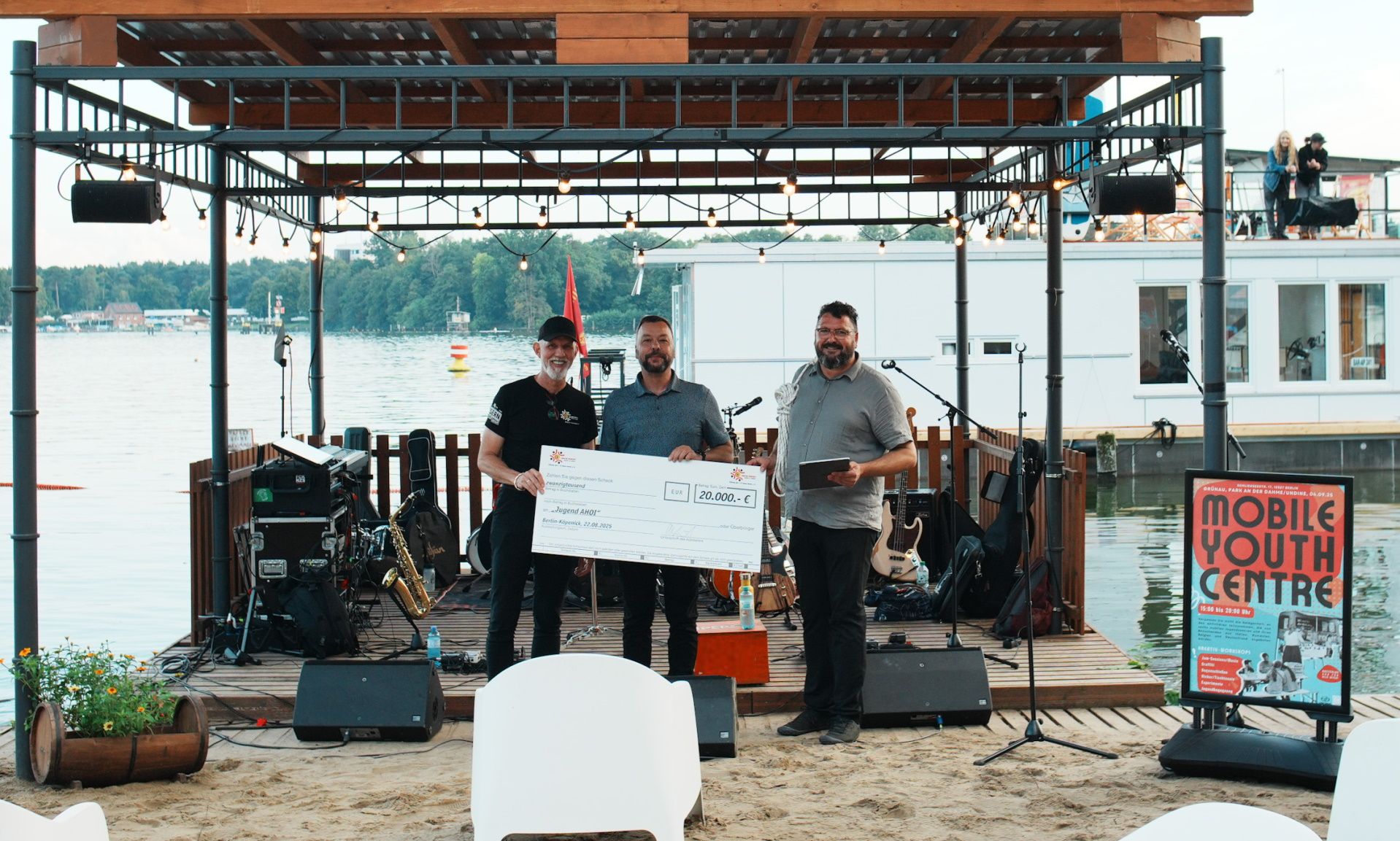 Drei Männer stehen auf einer Bühne am Wasser und halten einen großen Scheck. Im Hintergrund sind Musikgeräte und eine festliche Umgebung.