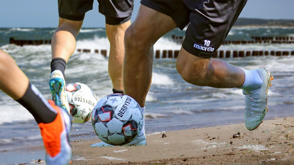 Players are dribbling a soccer ball on the beach while waves crash in the background.