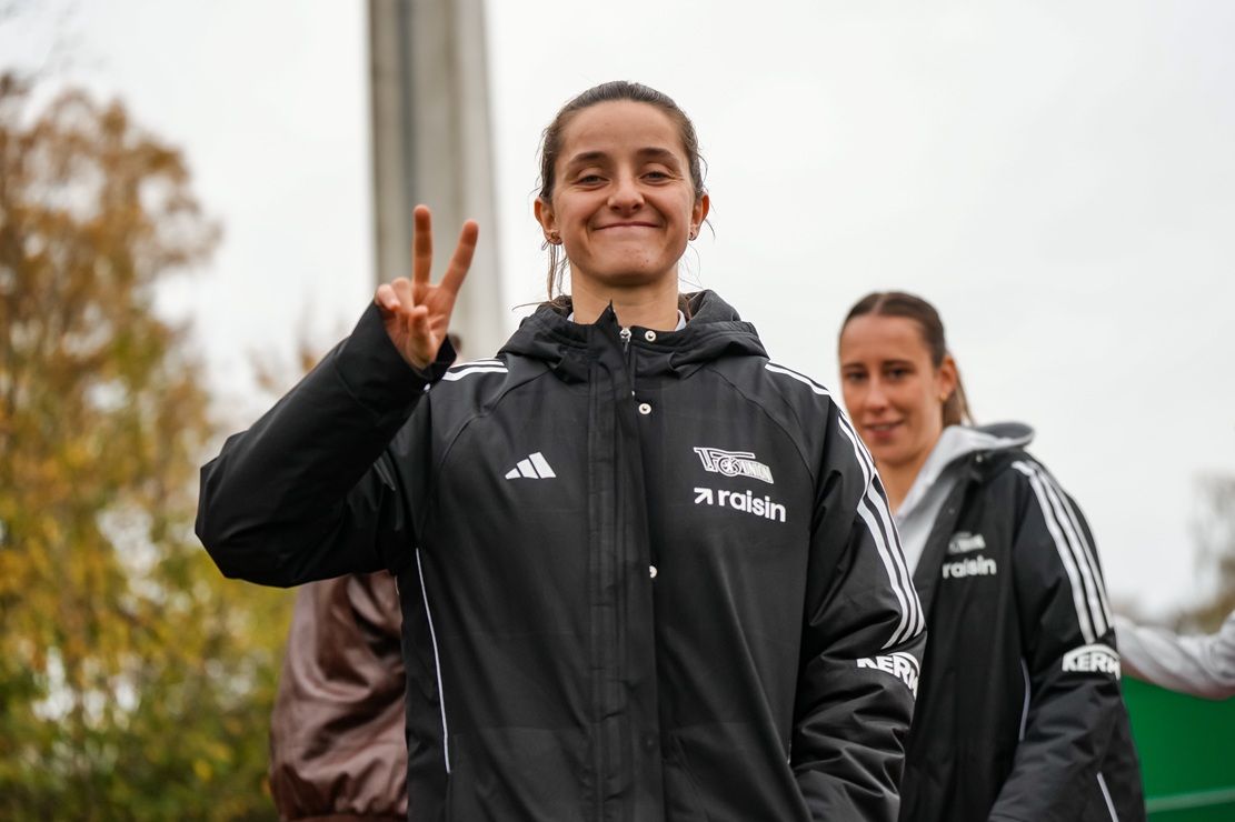Eine junge Frau in Sportkleidung lächelt und zeigt mit der Hand ein Peace-Zeichen. Im Hintergrund ist eine andere Person zu sehen.
