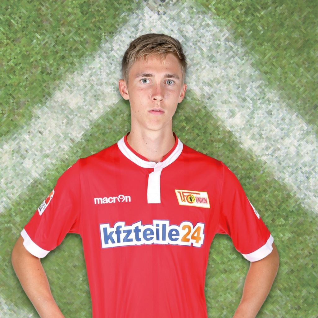 Ein junger Fußballspieler in einem roten Trikot mit einem Sponsorlogo steht vor einem grünen, texturierten Hintergrund.
