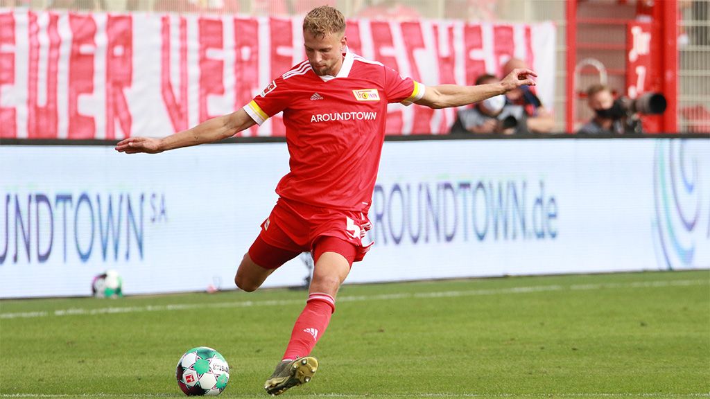 Ein Fußballspieler in rotem Trikot schießt den Ball während eines Spiels. Im Hintergrund sind Fans und Banner sichtbar.