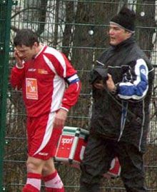 Ein verletzter Fußballspieler in rotem Trikot spricht am Telefon, während ein Betreuer in dunkler Kleidung ihn begleitet.