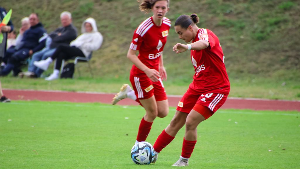 Eine Fußballspielerin in rotem Trikot schießt den Ball, während eine Teamkollegin in der Nähe steht. Zuschauer im Hintergrund.
