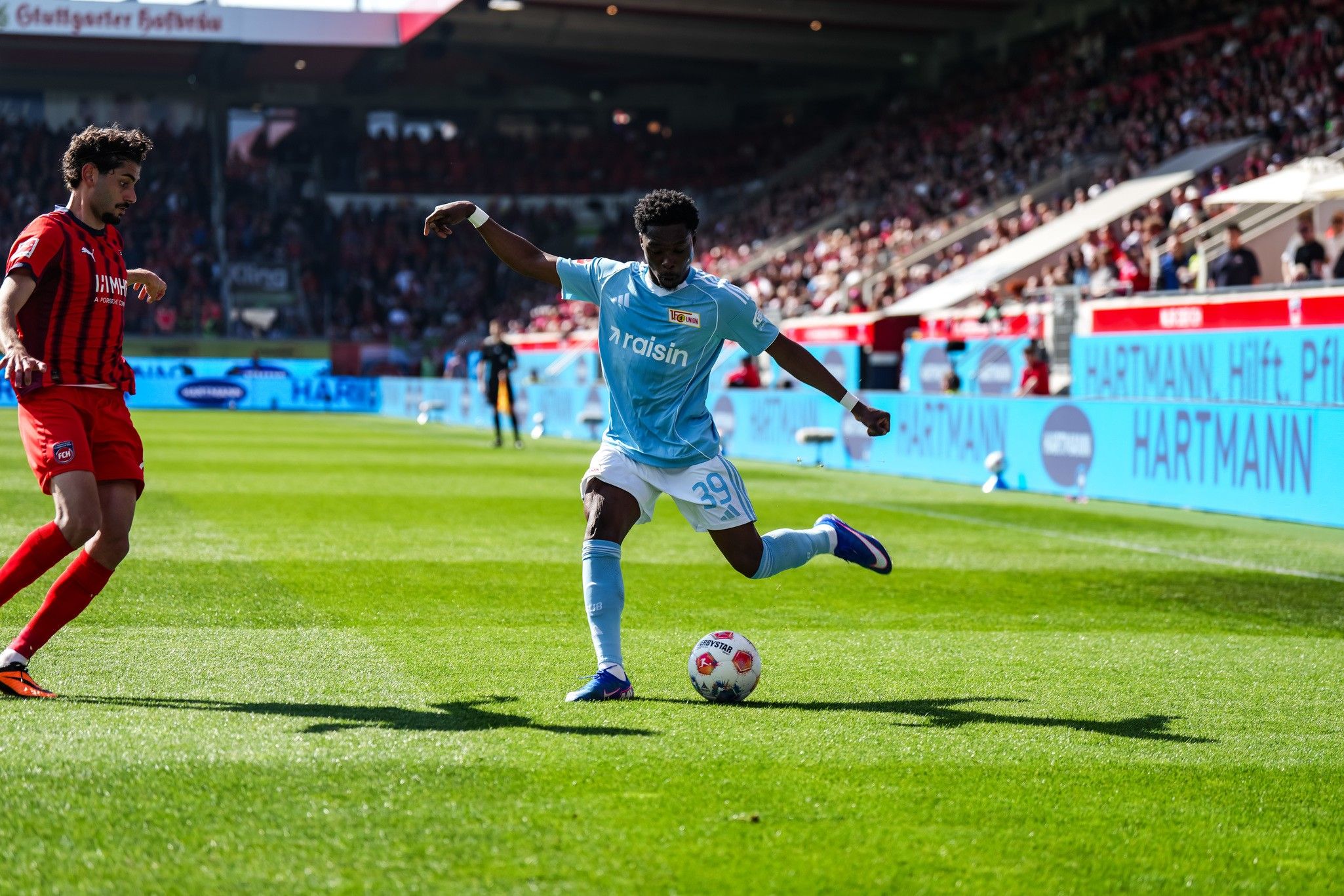 Spielszene im Stadion: Ein Spieler in blauem Trikot dribbelt den Ball, während ein Gegner in rotem Trikot ihn verfolgt.