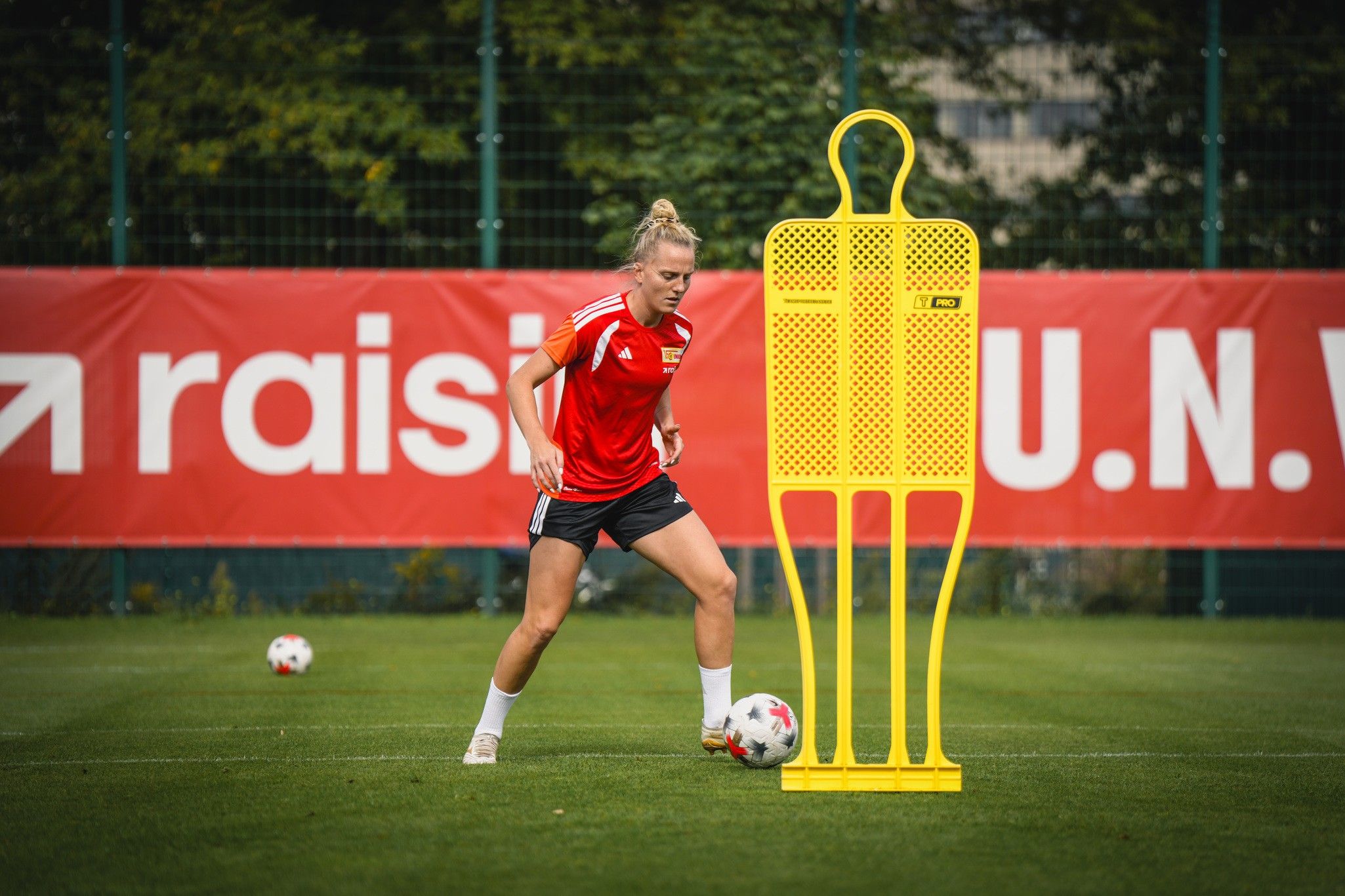 Eine Fußballspielerin in rotem Trikot trainiert im Freien, während sie um einen gelben Trainingsdummy dribbelt.