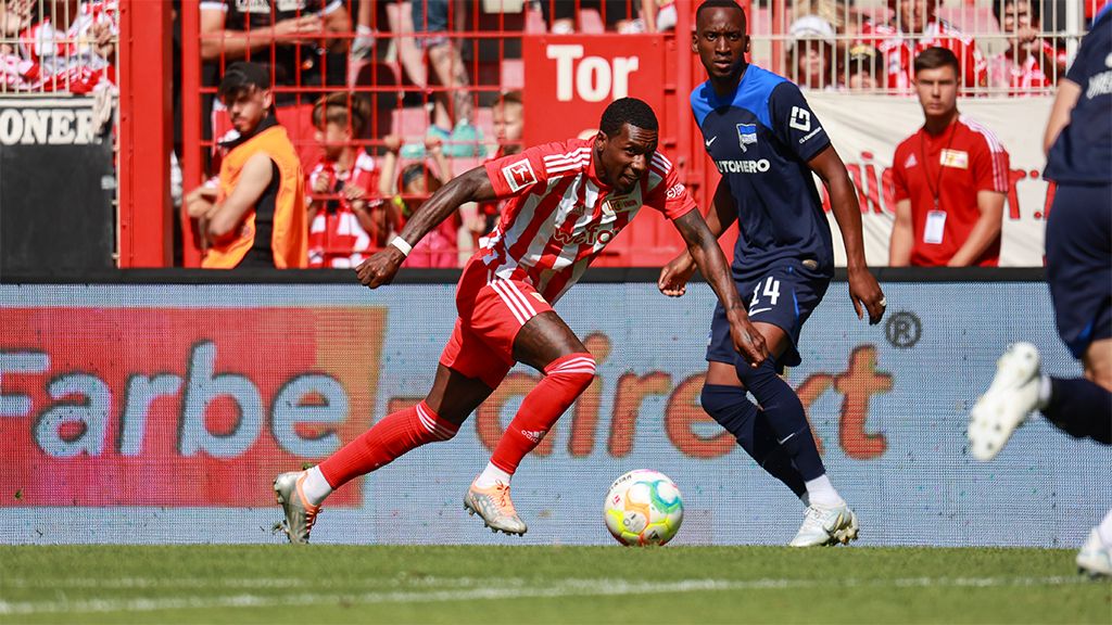 A football player in a red jersey dribbles the ball while an opposing player in a blue jersey chases him. Fans are visible in the background.
