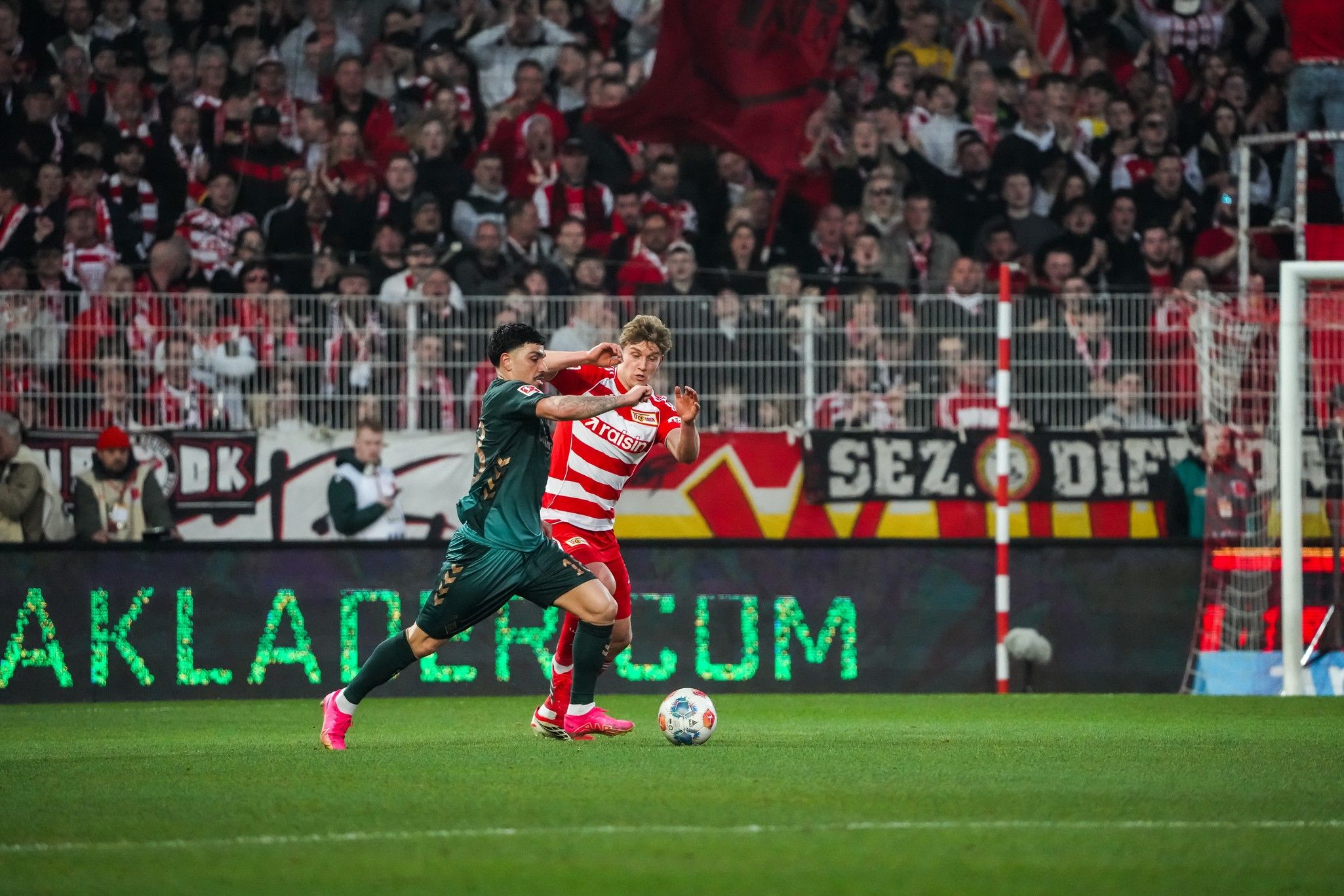 Ein Fußballspieler in roter und weißer Kleidung wird von einem gegnerischen Spieler abgehalten, während Zuschauer im Hintergrund jubeln.
